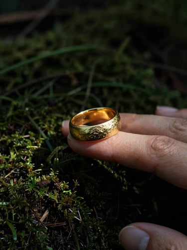 Moody editorial jewelry photography, close-up of a heavy textured gold ring on a finger resting on dark forest moss, high-contrast side light, deep shadows.