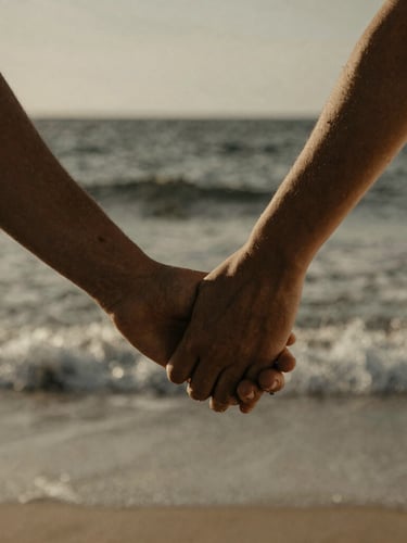 Close-up of a couple's hands gently intertwined, wind-blown ocean spray in the background, warm desaturated tones, dramatic natural light