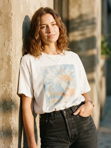 Medium-close candid shot of a model wearing a heavyweight white graphic t-shirt, leaning against a sun-drenched concrete wall, sharp shadows, warm golden hour light, 35mm film style