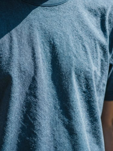Close-up of thick textured cotton fabric of a blue t-shirt, sunbeams casting sharp linear shadows across the weave, warm minimalist aesthetic