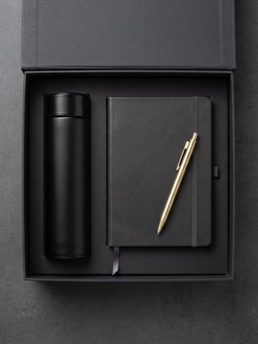 Crisp studio product photography, overhead flat-lay of a premium open corporate gift box containing a matte black thermos, leather notebook, and brass pen, deep charcoal and white tones, sharp focus, 85mm