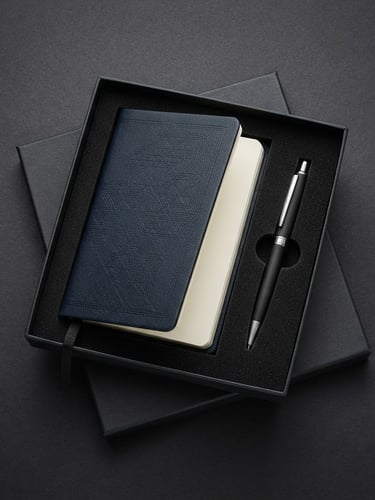 Crisp studio product photography of open premium corporate gift box on dark charcoal background, debossed leather notebook and metal pen visible, 85mm