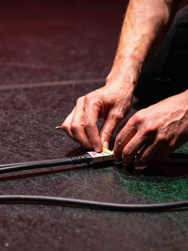 Close-up of a technician's hands carefully organizing and labeling thick black cables on a dark stage floor, warm spotlight from the side, sharp focus, 35mm