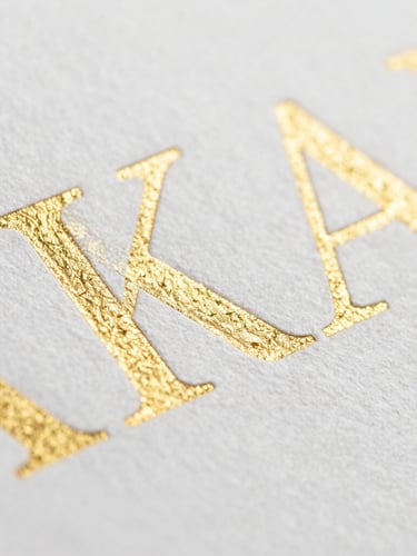 Macro close-up of heavy-weight textured paper with embossed gold foil lettering, crisp edges, bright clean studio lighting, shallow depth of field.