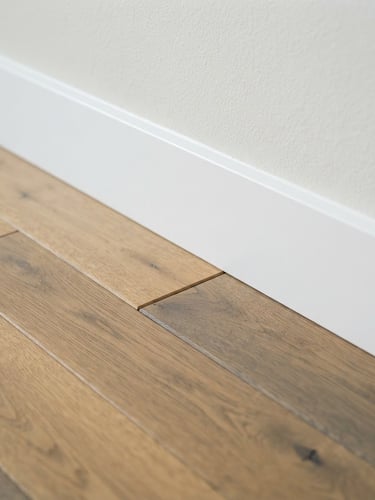 Macro architectural shot of perfectly aligned engineered hardwood planks meeting a clean white baseboard, bright natural coastal daylight, sharp focus, 35mm