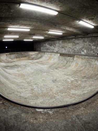 Wide panoramic shot of an empty indoor concrete skate bowl under raw overhead fluorescent lights, gritty texture, high contrast.