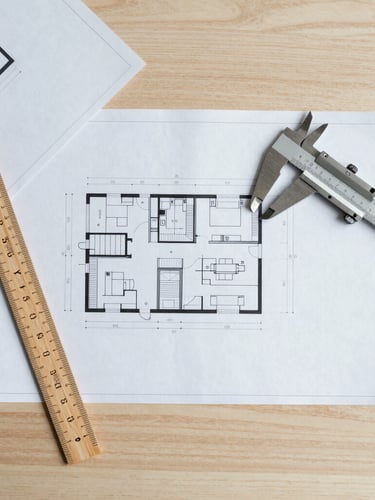 Overhead flat-lay of clean technical blueprints on a light oak desk, a wooden scale ruler and a metal caliper, warm diffused daylight
