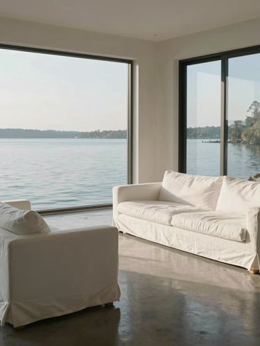 A sunlit, minimalist lakefront living room with white linen sofas, polished concrete floors reflecting soft north-facing light, and expansive windows looking out to quiet water.