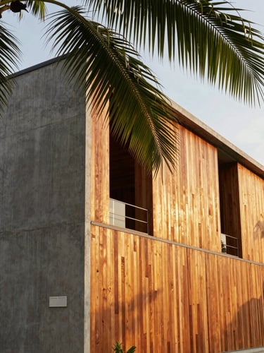 Architectural exterior of a modern tropical industrial house, raw concrete walls paired with warm teak wood panels, golden hour sunlight filtering through broad palm leaves, 35mm lens