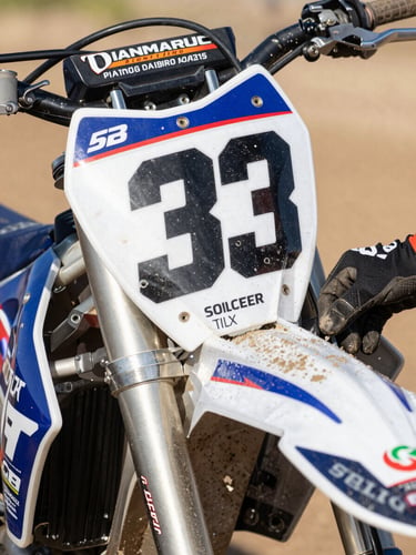 Close-up of a motocross bike fairing in the pit lane, sponsor decal sharp under direct afternoon sun, hands in mechanic gloves adjusting the number plate, dirt and oil visible on the panel surface