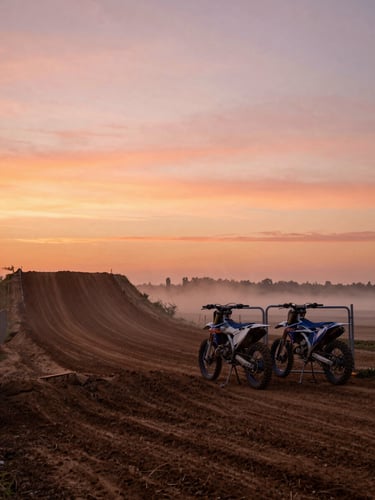 Wide panoramic view of a motocross track at dawn, starting gate and gate boards silhouetted against an orange-pink sky, mist rising from the dirt surface, empty track stretching to the left third, expansive sky dominating the upper frame