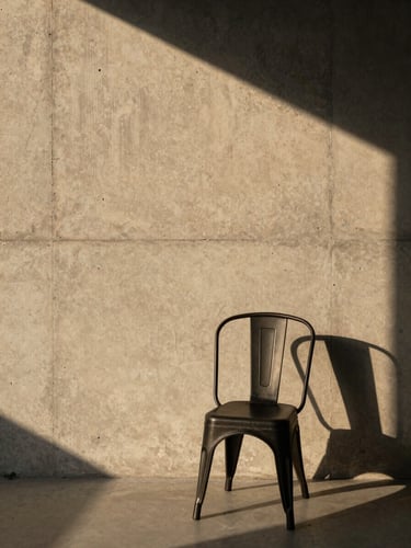 Straight-on elevation of a minimalist concrete wall with raw grain textures, a single black steel chair placed to the side, dramatic geometric shadows from natural golden hour light, high-contrast chiaroscuro.