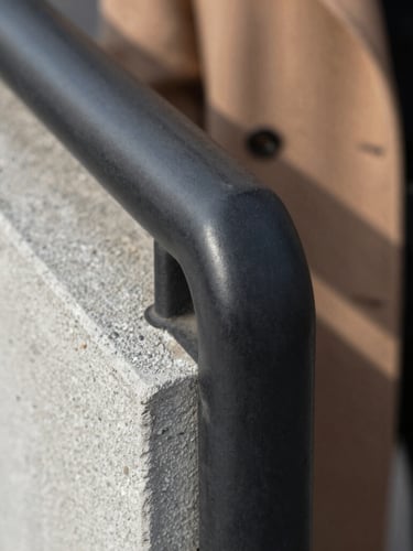 Extreme close-up of a matte black steel handrail meeting raw concrete grain, soft natural light highlighting the texture, a coat draped casually in the background.