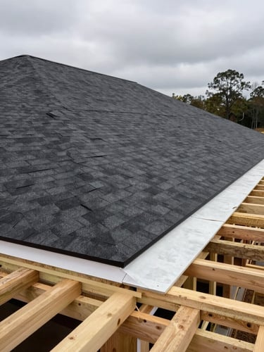 Wide environmental shot of a new construction residential build in Alabama, bare roof decking fully sheathed and crew laying the first course of dark shingles from the eave, overcast sky, raw lumber and felt paper visible in the foreground