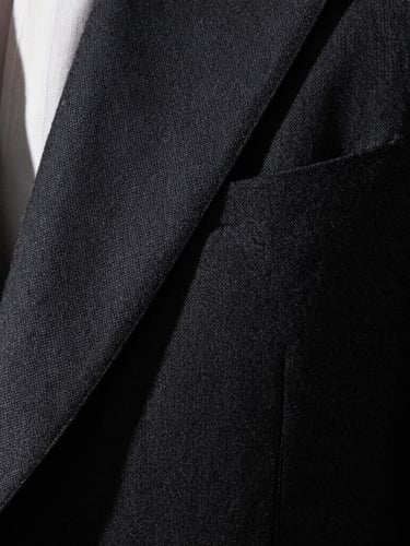 Extreme close-up of a dark tailored charcoal suit fabric, sharp focus on the weave, dramatic side lighting from a high window, deep shadows, gold cufflinks reflecting light.