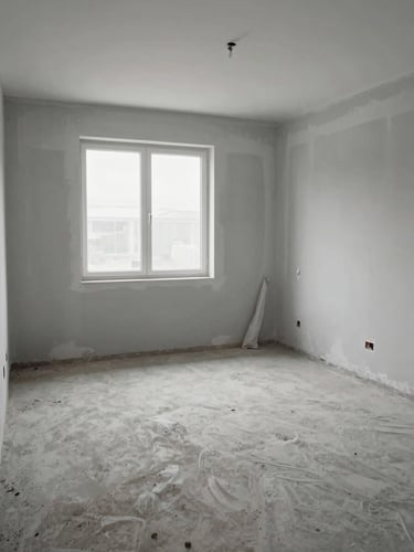 Wide shot of an empty room mid-renovation: bare concrete floor, one wall freshly plastered in light grey, diffused overcast daylight through an uncovered window, no people, dust sheets folded in corner, documentary style