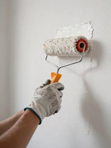 Close-up of a worker's hands in work gloves pressing a paint roller against a fresh white wall, soft north-facing daylight, paint texture visible at the edge of the stroke, no face visible, documentary framing