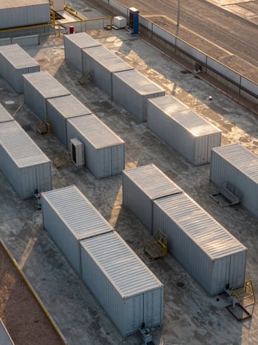 Overhead wide-angle view of a large battery storage container yard at an industrial plant in the UAE, containers arranged in rows on a concrete pad, late afternoon golden light casting long shadows across the steel units, plant infrastructure visible at the edges