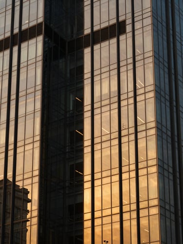 Architectural close-up of a modern corporate glass tower in Gurugram, dramatic golden hour light, deep charcoal shadows and warm gold reflections on the facade, sharp 35mm lens.