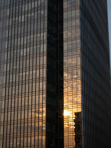 Dramatic architectural close-up of a modern glass skyscraper in Gurugram at golden hour, sharp reflections of light on steel and glass, deep charcoal tones with metallic gold highlights, sharp 35mm lens.