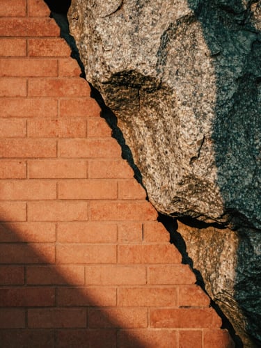 Close-up of earthen brickwork meeting a massive natural granite boulder, soft shadows, warm terracotta and dusty gray tones, golden hour light.
