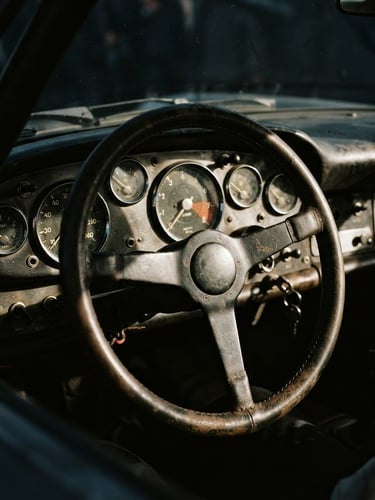 Close-up of a vintage racing car dashboard, mechanical dials with metal bezels, worn leather steering wheel, deep shadows, high-contrast natural light, 35mm film grain