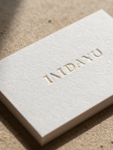 Macro shot of premium heavy-weight cotton business card with deep blind letterpress embossing and gold foil edge gilding, dramatic side lighting casting deep shadows on warm sand background.