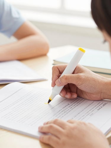 A tight close-up of a teacher's hand guiding a student's finger along a printed page, highlighting a word with a yellow marker, bright natural classroom light, shallow depth of field.