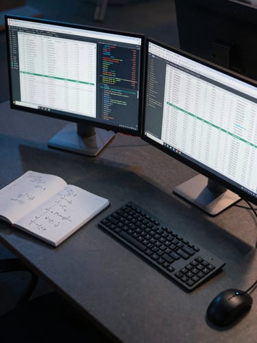 Overhead wide shot of a technical workstation under cool studio light, two monitors displaying dense financial data tables and code, a notebook with mathematical notation open to the left, sharp shadows on a dark desk surface