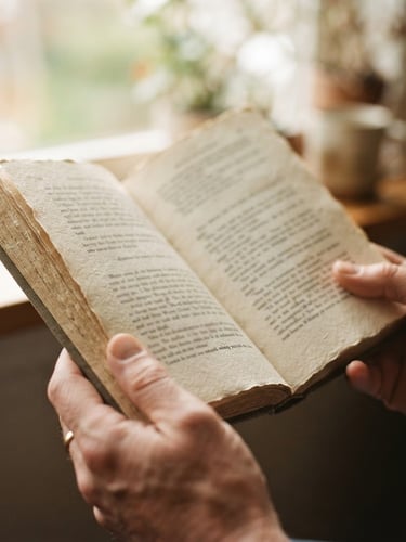 A close-up photograph of hands holding an open physical book with textured paper, warm morning window light illuminating the pages, shallow depth of field, 35mm style.