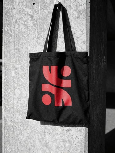 Low-angle shot of a heavy-duty black canvas tote bag with a bold red abstract graphic, slung over a shoulder against a stark concrete subway wall, harsh direct sunlight, gritty 35mm film texture