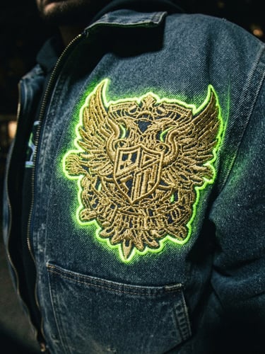 Close-up direct flash shot of heavy gold streetwear embroidery on a deep slate blue jacket, high-contrast neon under-glow
