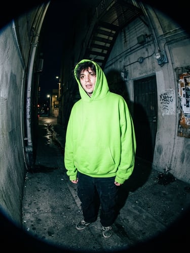 Gritty flash photography of a fan wearing an oversized electric lime green hoodie in a concrete alleyway, wide-angle fish-eye lens, direct lighting