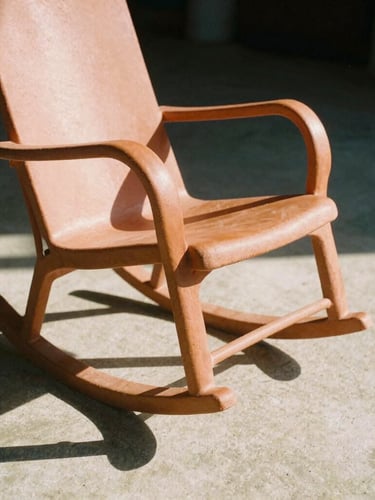 Low-angle sculptural shot of a terracotta rocking chair casting a sharp midday shadow on concrete, warm golden hour light, 35mm film