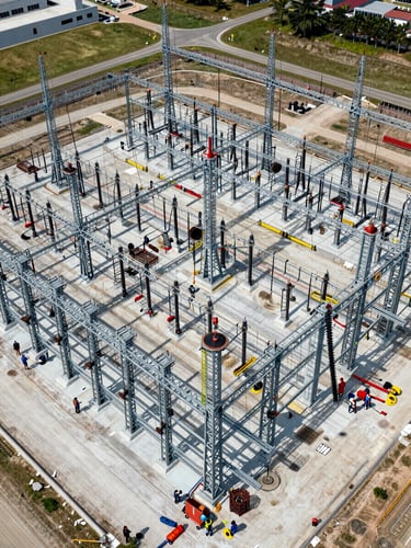 Wide overhead aerial view of a large substation under active construction, steel lattice structures being erected, crews coordinating on the ground below, midday industrial daylight, expansive site footprint visible