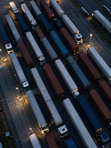 Overhead bird's-eye view of a busy freight logistics hub at dusk, dozens of trucks and containers arranged in systematic rows under cool floodlights, aerial perspective emphasizing scale and operational density, deep navy tones with silver-lit surfaces