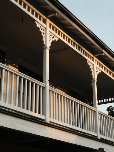 Tight architectural detail of a classic Brisbane Queenslander veranda, deep shadows under timber railings, warm golden hour light reflecting off white painted wood.