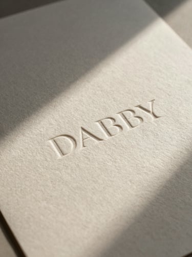 A high-angle portrait crop of physical debossed branding collateral on heavy handmade cotton paper, soft north-facing daylight casting elegant shadows, minimalist composition.
