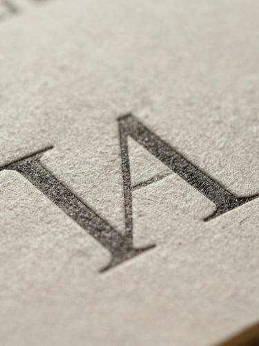 Extreme macro crop of heavy cotton paper stock with deep debossed typography under soft, diffused north daylight, showing rich tactile texture.