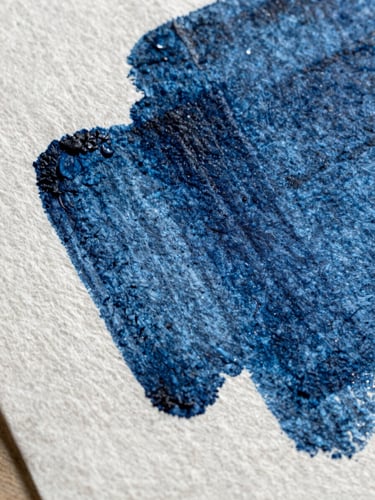 Extreme close-up of heavy-weight textured cotton paper with deep indigo ink pressed into the grain, casting a sharp shadow across the surface under harsh natural light.