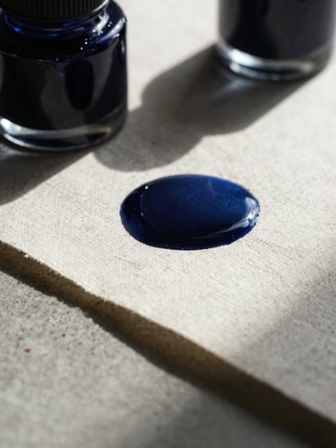 Extreme close-up macro shot of textured raw linen paper and deep indigo ink wells on a concrete table, dramatic hard natural light casting long sharp shadows.