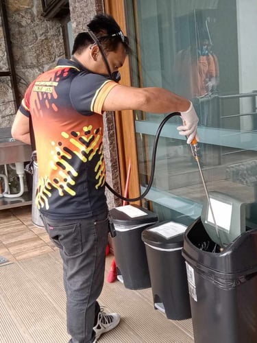 Professional pest control technician spraying disinfectant into outdoor trash bins using a pressure wand.