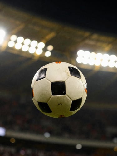 Dynamic close-up of a spinning football on a dark pitch under glowing gold stadium floodlights, dramatic sports photography, high-contrast backlighting, 35mm