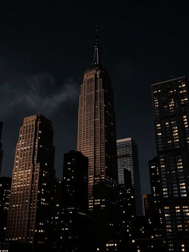 Low-angle cinematic shot of the NYC skyline at midnight, high-contrast chiaroscuro, deep shadows with subtle golden rim lighting on skyscrapers, atmospheric smoke haze, anamorphic lens