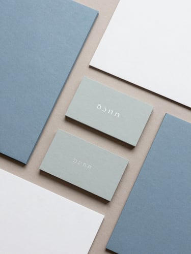 Top-down flat lay of coordinated brand collateral, soft slate blue and muted sage business cards, clean stationery aligned at 90-degree angles on a warm taupe surface, soft diffused overcast daylight, 35mm
