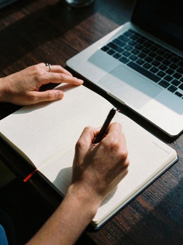 High-contrast close-up of a creator's hands writing notes in a physical journal next to an open laptop, harsh afternoon sunlight casting deep shadows, 35mm film grain.