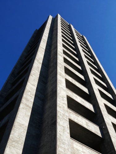 High-contrast wide-angle editorial photography of a concrete brutalist tower under direct harsh sunlight, deep black shadows, sharp angles, vibrant blue sky.