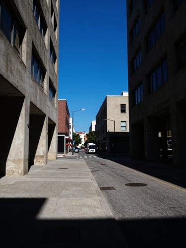 High-contrast urban street scene with sharp shadows and direct sunlight, concrete architecture, bold blue sky