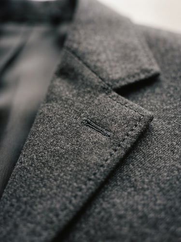 Macro studio photograph of a structured charcoal suit jacket lapel, soft natural window light, highlighting premium wool texture and clean hand-finished stitching, 35mm