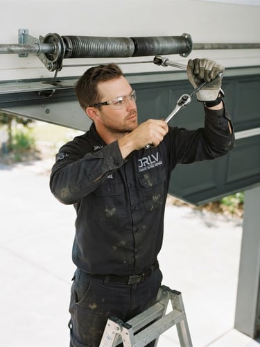 Close-up of a JRLV Garage Doors Repairs technician in branded uniform, safely working on a garage door spring, bright natural outdoor daylight, clean steel and charcoal tones, sharp focus, 35mm.